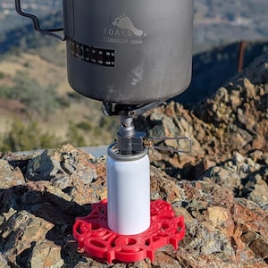 Puede incluir: Un termo de camping de titanio negro con asa, sobre una estufa portátil y una base roja. El termo tiene el texto "TOAKS TITANIUM 750ml". El conjunto está sobre una superficie rocosa, al aire libre.