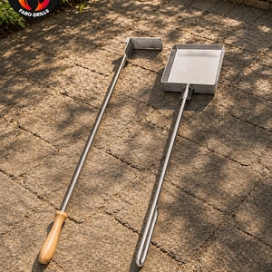 May include: Two stainless steel grill tools featuring long handles. One tool is a flat-edged scraper with a wooden handle, and the other is a rectangular ash shovel with a looped metal handle. The logo FABO GRILLS with a flame icon is visible.