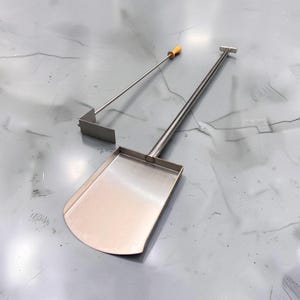 May include: A stainless steel pizza peel with a long handle and a flat, rectangular blade with rounded edges, accompanied by a separate metal tool featuring a thin rod, a small wooden handle, and a perpendicular rectangular scraper attachment.