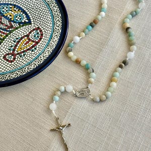Op de afbeelding: Een rozenkrans met veelkleurige edelsteen kralen, een zilveren kruisbeeld en een zilveren middelpunt. De kralen zijn in tinten wit, blauw, groen en bruin. Een decoratieve schaal met een blauwe rand en mozaïek visontwerp staat op de achtergrond.