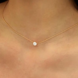 Può includere: Una delicata collana d'oro con un singolo pendente rotondo e trasparente. Il pendente è centrato su una sottile catena d'oro, creando un design minimalista ed elegante. La collana è mostrata su uno sfondo color carne.