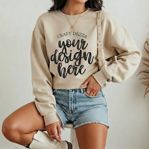 Puede incluir: Una sudadera beige con el texto "Craft Dazza your design here" en escritura negra. La modelo lleva pantalones cortos vaqueros y botas de vaquero blancas. La sudadera tiene mangas largas y cuello redondo.