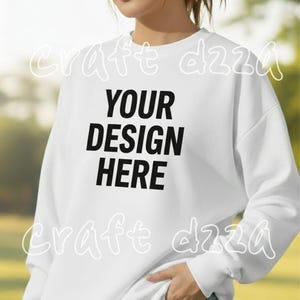 Puede incluir: Sudadera blanca con el texto "YOUR DESIGN HERE" en letras negras. La sudadera tiene mangas largas y cuello redondo. La modelo lleva vaqueros azules.