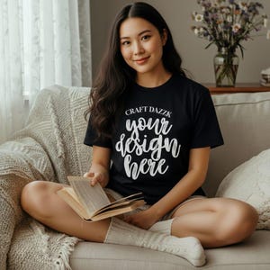 Puede incluir: Camiseta negra con el texto "Craft Dazzk your design here" en escritura blanca. La persona está sentada en un sofá y sosteniendo un libro. La persona lleva calcetines blancos.