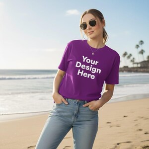 Puede incluir: Una camiseta morada con el texto "Your Design Here" en blanco. La camiseta la lleva una persona en una playa. La persona lleva gafas de sol y vaqueros azules.