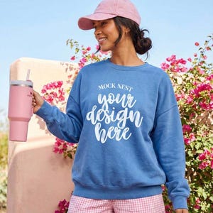 Puede incluir: Una mujer con una gorra de béisbol rosa, una sudadera azul con el texto "MOCK NEST your design here" y pantalones cortos de cuadros rosas. Sostiene un vaso térmico rosa con una pajita. Flores de buganvillas en el fondo.