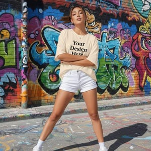 Puede incluir: Una persona con una camiseta color crema y pantalones cortos blancos se encuentra frente a una pared de ladrillos cubierta de grafitis. La camiseta tiene el texto "Your Design Here" impreso. La persona lleva zapatillas blancas.