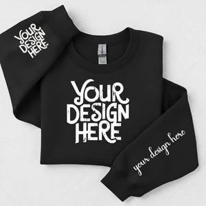 Puede incluir: Sudadera negra de cuello redondo con el texto "YOUR DESIGN HERE" en blanco en la parte delantera y las mangas. Las mangas también presentan el texto "your design here" en letra cursiva. La sudadera está doblada.