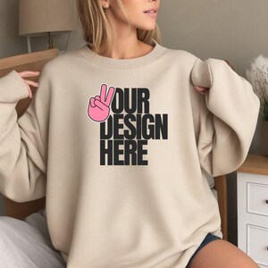 Puede incluir: Sudadera beige con el texto negro "YOUR DESIGN HERE", acompañada de un signo de la paz rosa. Esta prenda de cuello redondo y manga larga es ideal para el uso diario.