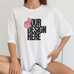 Puede incluir: Camiseta blanca con un signo de la paz rosa y el texto negro "YOUR DESIGN HERE". La camiseta tiene mangas cortas y un corte holgado.