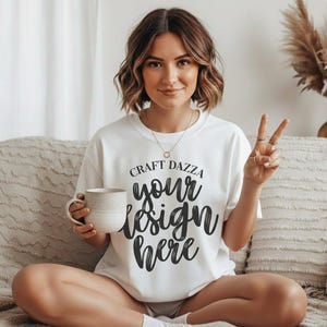 Puede incluir: Camiseta blanca con el texto "CRAFT DAZZA your design here" en escritura negra. La persona sostiene una taza beige y hace un signo de paz. La persona está sentada en un sofá beige.