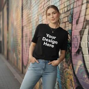 Puede incluir: Camiseta negra con el texto blanco "Your Design Here". La camiseta es de cuello redondo. La persona lleva vaqueros azules y está de pie frente a una pared de ladrillos con graffiti.