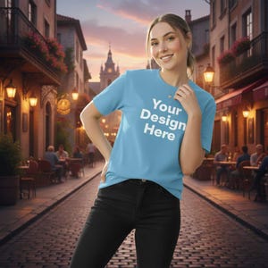 Puede incluir: Una camiseta azul claro con el texto "Your Design Here" en blanco. La camiseta se usa con pantalones negros. El fondo muestra una escena de calle europea con edificios y asientos al aire libre.