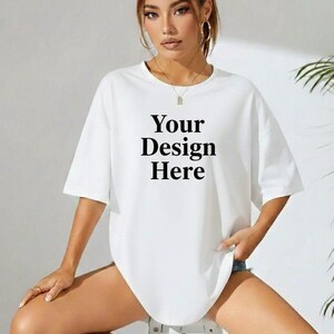 Puede incluir: Camiseta blanca de gran tamaño con el texto "Your Design Here" en negrita negra. La camiseta tiene mangas cortas y un corte holgado. La modelo lleva la camiseta y está sentada en un taburete.