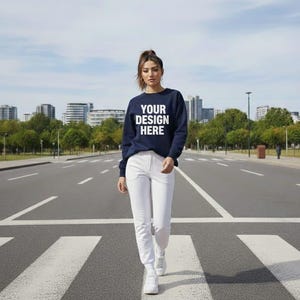 Puede incluir: Una persona lleva una sudadera azul marino con el texto "YOUR DESIGN HERE" en blanco, combinada con pantalones blancos y zapatillas deportivas. La persona camina por un paso de cebra en un entorno urbano.
