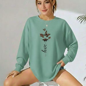 Puede incluir: Sudadera verde claro con un diseño de mariposas y la palabra "love" en cursiva. Las mariposas tienen patrones marrones y negros. La sudadera tiene mangas largas y cuello redondo.