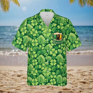 May include: Green short-sleeved shirt with a pattern of shamrocks and leaves. The shirt features a collar and buttons. A graphic of three beer mugs with the word "Sl&aacute;inte" is on the front.