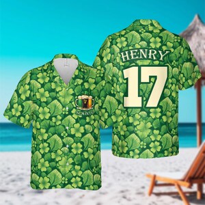 May include: Green short-sleeved shirt with a pattern of shamrocks and leaves. The shirt has a V-neck and features the name "HENRY" and the number "17" on the back. The front of the shirt has a graphic of beer mugs and the word "Slainte".