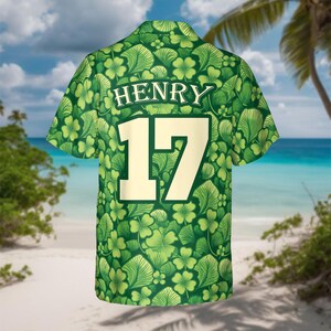 May include: Green short-sleeved shirt with a pattern of shamrocks and leaves. The name "HENRY" is printed above the number "17" in a cream color. The shirt is set against a beach background with palm trees and blue water.