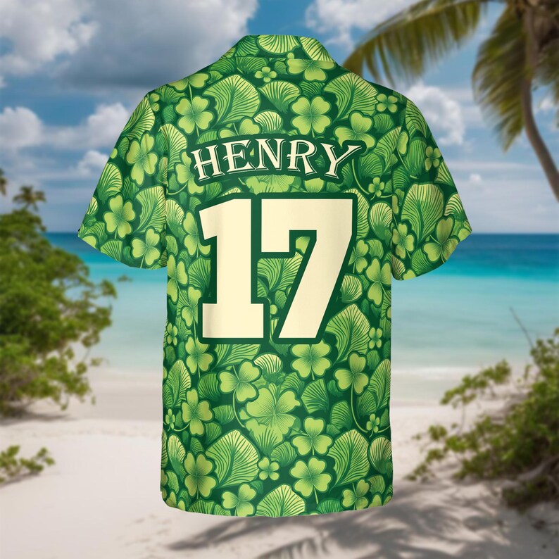 May include: Green short-sleeved shirt with a pattern of shamrocks and leaves. The name "HENRY" is printed above the number "17" in a cream color. The shirt is set against a beach background with palm trees and blue water.