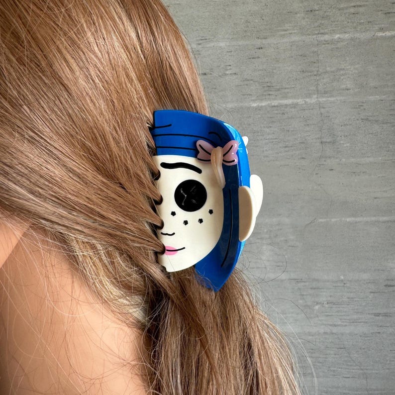 May include: A novelty hair clip in the shape of a cartoon face, with blue hair, a pink bow, and black button eyes. The clip is attached to brown hair, offering a distinctive hair accessory.