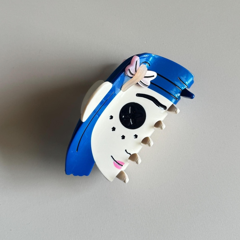 May include: A blue and white hair claw clip with a cartoon face design. The clip has a blue top with white accents, a black button eye, pink lips, and a small pink flower. The clip is made of plastic.
