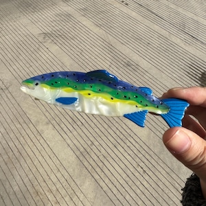 Op de afbeelding: Een decoratief visvormig object met heldere kleuren. De vis heeft een blauw, groen en geel patroon met zwarte stippen. De staart en vinnen zijn helderblauw. De vis wordt door een hand vastgehouden tegen een betonnen achtergrond.