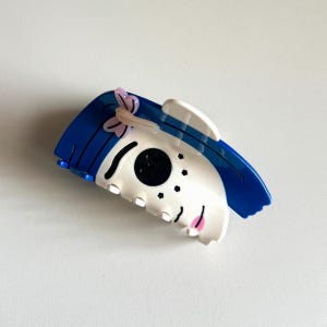 May include: A blue and white hair claw clip with a cartoon face design. The clip features blue hair, a pink bow, and a white face with black and pink details. The clip is made of plastic and has a claw-like design.