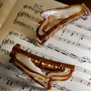 May include: Two brown and white hair clips with a decorative design, resting on sheet music. The clips have a tortoiseshell pattern and are embellished with small, clear rhinestones. The sheet music has musical notes and the text "Var. II Brillante".