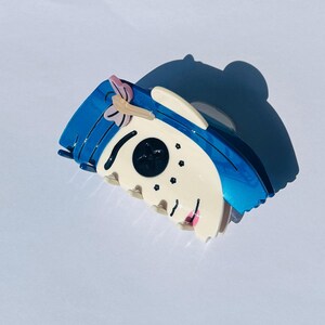 May include: A blue and white hair claw clip with a cartoon face design. The clip has a blue top with a pink bow, a white face, and black eyes. The clip is made of a shiny material.