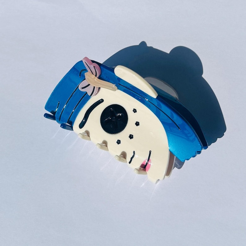 May include: A blue and white hair claw clip with a cartoon face design. The clip has a blue top with a pink bow, a white face, and black eyes. The clip is made of a shiny material.