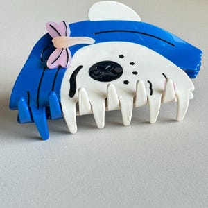 May include: A blue and white hair claw clip with a decorative butterfly accent. The clip has a shark-like design with a white skull-shaped centre and blue accents. The clip has multiple teeth for securing hair.
