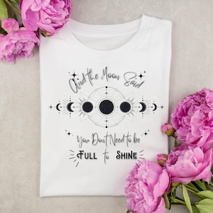 Puede incluir: Camiseta blanca con un diseño celestial y el texto "And the Moon Said, You Don't Need to be FULL to SHINE." El diseño presenta lunas, estrellas y formas geométricas. Peonías rosas en el fondo.