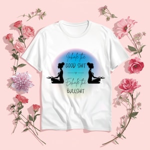 May include: White t-shirt with the text "Inhale the GOOD SHIT, Exhale the BULLSHIT" in a script font. The design includes two black silhouettes of people in a yoga pose, set against a gradient background. Pink flowers surround the shirt.