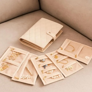 May include: A cream-colored jewelry organizer with a quilted cover and snap closure, alongside several open pouches. The pouches, also cream-colored, feature clear pockets and gold zippers, displaying various necklaces, bracelets, and earrings.