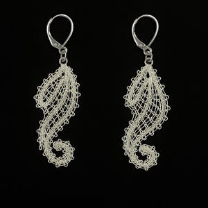 May include: A pair of silver-colored earrings in a seahorse design. The earrings are made of a delicate, open-weave material, with a curved shape and a small loop at the top for the ear wire. The background is black.