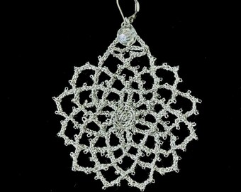 Può includere: Un orecchino fatto a mano di colore argento con un delicato design traforato. L'orecchino presenta una forma a goccia con un motivo floreale centrale e un accento di cristallo scintillante nella parte superiore. L'intricato motivo a forma di pizzo è visibile su sfondo nero.