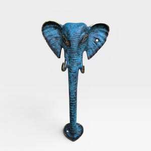 May include: A metal door handle shaped like an elephant head with a long trunk extending downward. The surface features a textured, weathered blue patina finish with dark bronze undertones visible on the ears, tusks, and trunk details.