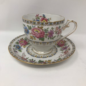 May include: A white teacup and saucer set with a floral design. The cup and saucer feature pink, blue, and purple flowers with gold trim. The teacup has a curved handle and a scalloped rim.