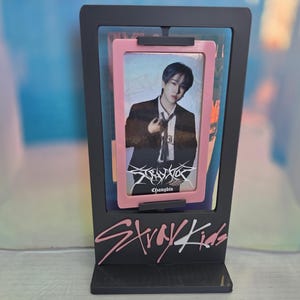 Puede incluir: Un expositor negro con una tarjeta fotográfica de una persona con traje, enmarcada en rosa. El expositor tiene el texto "Stray Kids" en letras rosas y blancas.