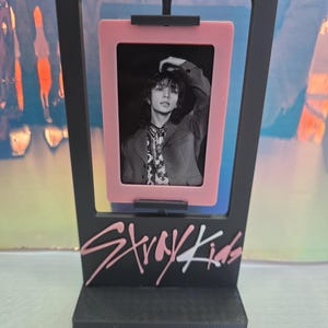 Puede incluir: Un marco de fotos negro con un marco interior rosa que contiene una foto en blanco y negro. El marco tiene la inscripción rosa "Stray Kids" en la parte inferior.