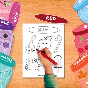 Puede incluir: Una mano infantil colorea una p&aacute;gina con un rotulador rojo. La p&aacute;gina presenta dibujos lineales de una cereza, una manzana y un bast&oacute;n de caramelo, con la palabra "RED" en la parte superior. Frascos coloridos con etiquetas de colores rodean la p&aacute;gina.