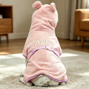 Könnte beinhalten: Ein rosa Hunde-Hoodie mit Bärenohren, einem lila Geschirr und dem Namen "Mila" auf dem Rücken gestickt. Der Hund sitzt auf einem hellen Teppich. Der Hoodie hat eine weiche, plüschige Textur.