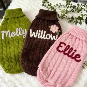 Pode incluir: Três camisolas para cães de malha entrançada em verde, castanho e rosa. Cada camisola tem um nome diferente bordado: "Molly", "Willow" e "Ellie". A camisola castanha tem uma pequena aplicação floral.