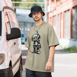 May include: A beige oversized t-shirt featuring a cartoon robot design. The robot is gray and black with yellow eyes and is wearing a white shirt and black sneakers. The person in the image is wearing a black bucket hat and a silver chain.