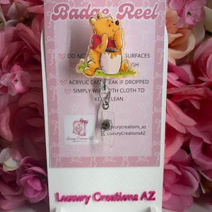 May include: A badge reel featuring a yellow bear character wearing a red shirt and holding a honey pot. The badge reel is attached to a pink display card with the text Badge Reel and Luxury Creations AZ, providing care instructions for the acrylic item.