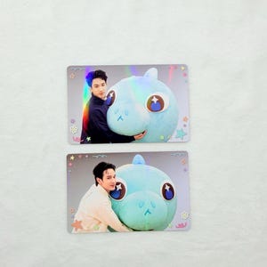 Peut inclure: Deux cartes à collectionner rectangulaires montrant une personne serrant dans ses bras une grande créature en peluche bleu clair aux yeux ronds. Les cartes ont une finition holographique et sont ornées de petites icônes colorées comme des étoiles.