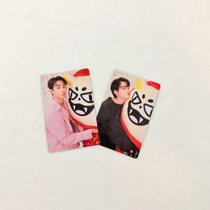 May include: Two rectangular photo cards featuring a person posing with a cartoon character. The left card shows a person in a pink shirt, while the right card shows a person in a black shirt. Both cards feature a white, horned character with a wide grin.