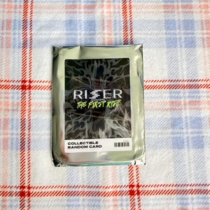 May include: A sealed collectible card pack with the text "RISER CONCERT THE FIRST RISE" and "COLLECTIBLE RANDOM CARD" on a white label. The pack is silver and sits on a red, white, and blue plaid fabric.