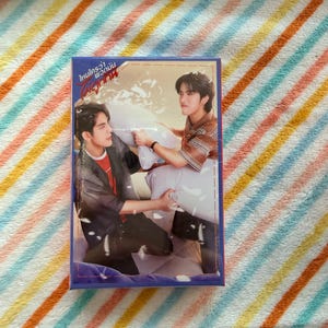 Può includere: Una scatola rettangolare con bordo blu raffigurante due uomini in posa romantica. La scatola ha un testo in lingua straniera. Lo sfondo è un motivo a strisce colorate.